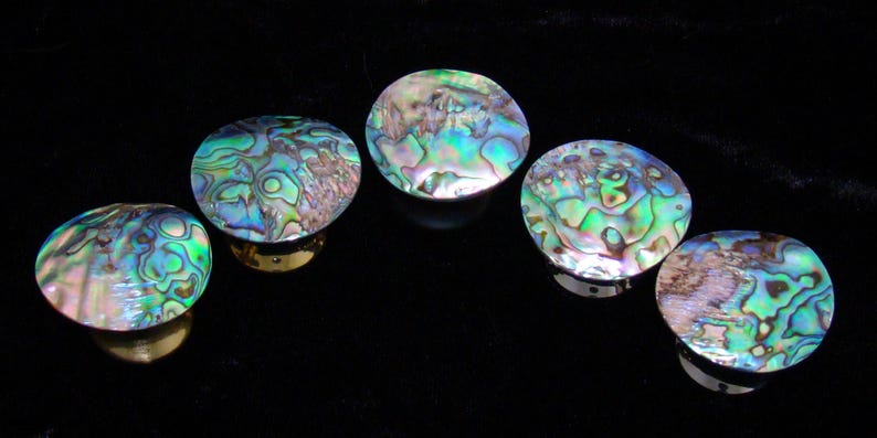May include: Five circular cabinet knobs featuring iridescent abalone shell inlays with swirling patterns of green, blue, and purple. Each knob has a polished metallic base that supports the rounded, shimmering shell top, creating a reflective surface.