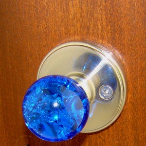 May include: A close-up of a door knob with a bright blue, cracked glass sphere. The knob is set against a shiny, silver-colored circular plate, mounted on a wooden door. The door has a warm, brown tone.