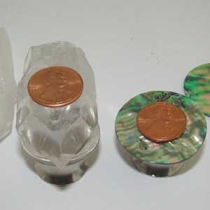 May include: Clear quartz crystal and two decorative bottle stoppers with abalone shell tops. Each stopper has a copper penny on top. The abalone shell has iridescent green, blue, and purple hues.