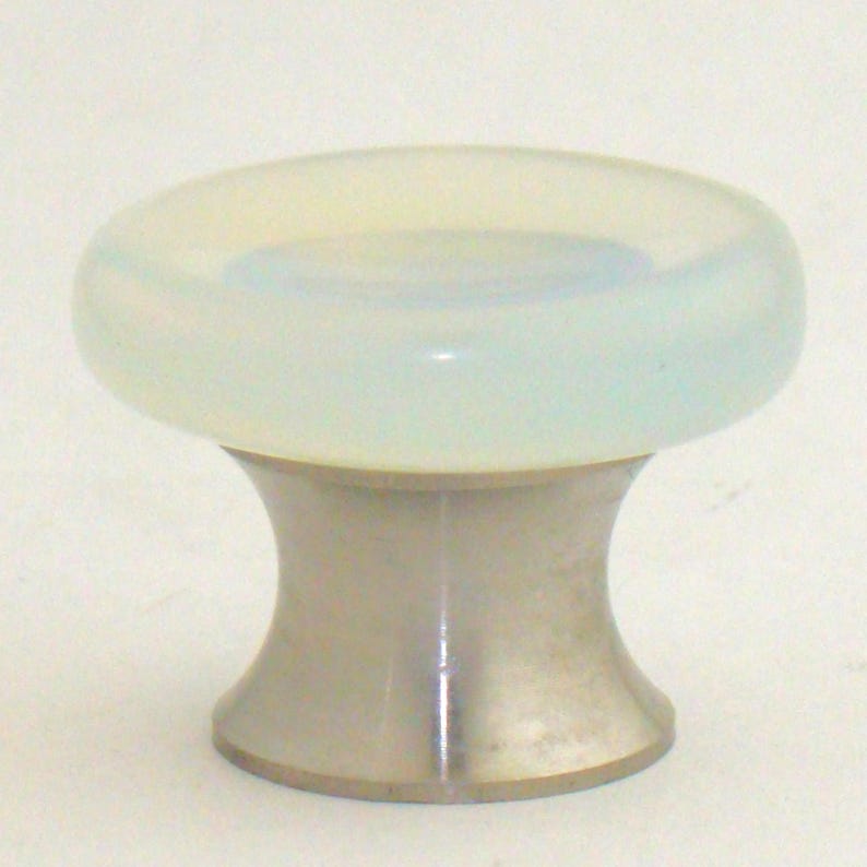 K&ouml;nnte beinhalten: Ein M&ouml;belknopf mit einer glatten, durchscheinenden, bl&auml;ulich-wei&szlig;en runden Oberseite mit sanftem opaleszierendem Glanz. Der Sockel besteht aus poliertem Metall und weist eine ausgestellte, konkave Form auf, die sich zur Mitte hin verj&uuml;ngt.