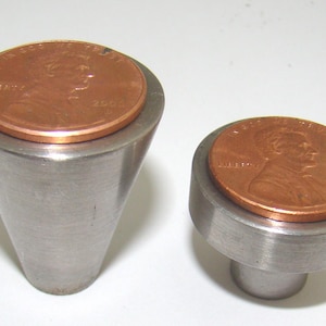 May include: Two metal cabinet knobs with a copper penny on top. One knob is cone-shaped, the other is cylindrical. The knobs are made of brushed metal and are set against a white background. The pennies are dated 2000.