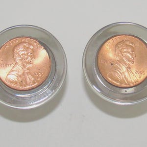 May include: Two clear, circular coin holders each containing a copper penny. The pennies are dated 2000 and 2006, and feature the profile of Abraham Lincoln. The holders are on a light background.