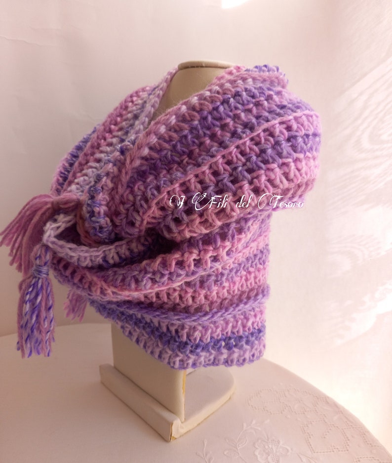 May include: A purple and pink crocheted shawl with a fringe tassel. The shawl is displayed on a white mannequin.