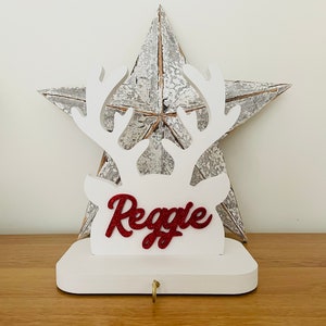 May include: A white wooden Christmas stocking holder shaped like a reindeer head with antlers. The name "Reggie" is written in red glitter. A gold hook is at the bottom. A rustic metal star is in the background.