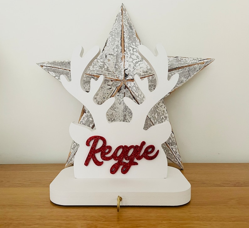 May include: A white wooden Christmas stocking holder shaped like a reindeer head with antlers. The name "Reggie" is written in red glitter. A gold hook is at the bottom. A rustic metal star is in the background.