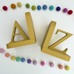 a pair of letter bookends, A & Z, painted in a mustard colour, on a white background with two strings of coloured felt balls used as a photo prop