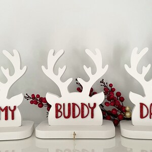 May include: Three white wooden reindeer head decorations with red glitter text that reads "MUMMY", "BUDDY", and "DADDY". Each reindeer has a small hook below the name. Red berries are placed between the reindeer.