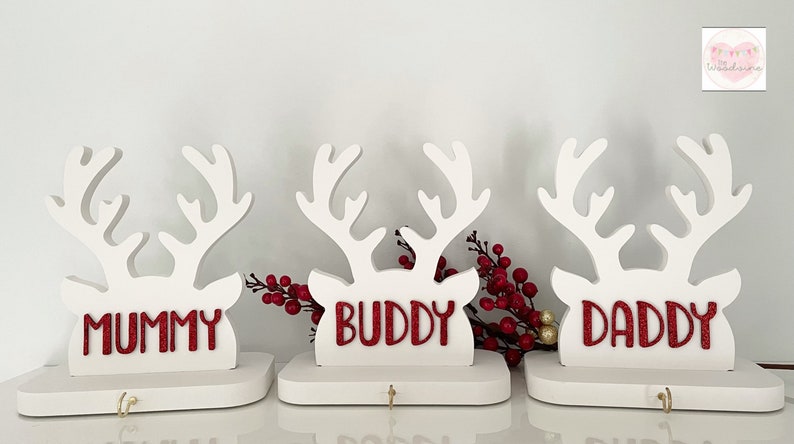 May include: Three white wooden reindeer head decorations with red glitter text that reads "MUMMY", "BUDDY", and "DADDY". Each reindeer has a small hook below the name. Red berries are placed between the reindeer.