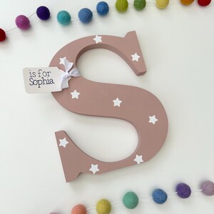 Pode incluir: Uma grande letra "S" de madeira rosa empoeirado decorada com estrelas brancas. Uma pequena etiqueta amarrada com uma fita branca diz "is for Sophia". Uma guirlanda de bolas de feltro coloridas &eacute; pendurada acima e abaixo da letra. Perfeito para um quarto infantil.