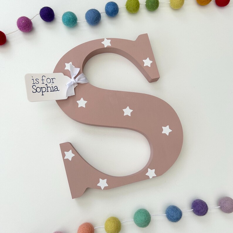 Pode incluir: Uma grande letra "S" de madeira rosa empoeirado decorada com estrelas brancas. Uma pequena etiqueta amarrada com uma fita branca diz "is for Sophia". Uma guirlanda de bolas de feltro coloridas &eacute; pendurada acima e abaixo da letra. Perfeito para um quarto infantil.