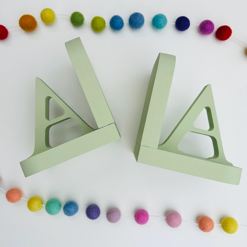 a pair of letter bookends, A & A, painted in a soft green colour, on a white background with two strings of coloured felt balls used as a photo prop