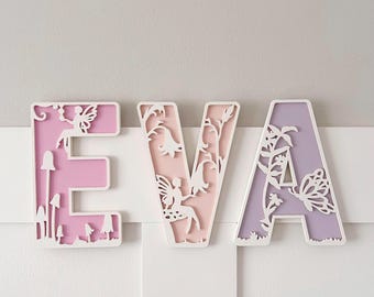 May include: Three-dimensional wooden letters spelling "EVA" in white, pink, and lavender. Each letter features intricate cut-out designs of fairies, flowers, and mushrooms. The letters are mounted on a white surface.