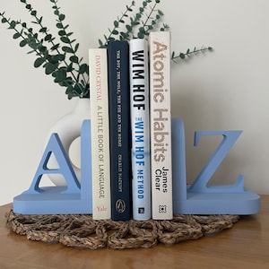 A pair of blue painted wooden bookends, shaped as the letters 'A' and 'Z’, supporting a book, with a white vase containing artificial greenery visible in the background, all on a woven surface