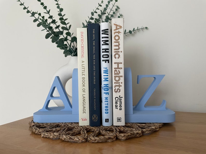 A pair of blue painted wooden bookends, shaped as the letters 'A' and 'Z’, supporting a book, with a white vase containing artificial greenery visible in the background, all on a woven surface