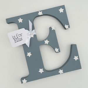 Pode incluir: Uma letra "E" cinzenta de madeira decorada com estrelas brancas. Uma etiqueta branca amarrada com uma fita branca diz "is for Ellis". A letra &eacute; um item decorativo, possivelmente para um ber&ccedil;&aacute;rio ou quarto infantil.