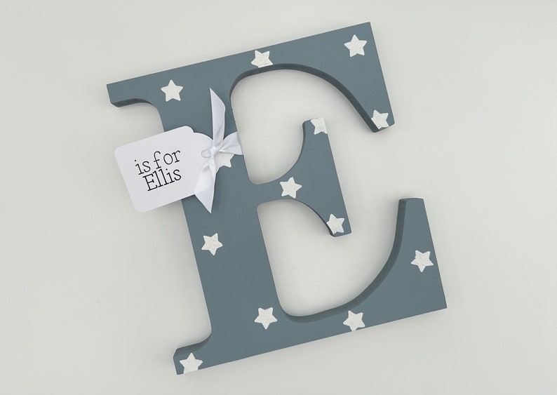 Pode incluir: Uma letra "E" cinzenta de madeira decorada com estrelas brancas. Uma etiqueta branca amarrada com uma fita branca diz "is for Ellis". A letra &eacute; um item decorativo, possivelmente para um ber&ccedil;&aacute;rio ou quarto infantil.