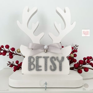 May include: White wooden reindeer head with antlers, a gray ribbon, and the name "BETSY" in silver glitter letters. A small silver bell and a hook are attached. Red berries and gold ornaments decorate the background. A festive Christmas decoration.