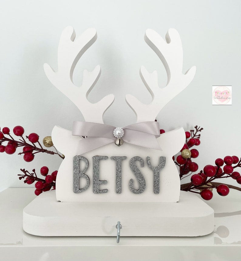 May include: White wooden reindeer head with antlers, a gray ribbon, and the name "BETSY" in silver glitter letters. A small silver bell and a hook are attached. Red berries and gold ornaments decorate the background. A festive Christmas decoration.
