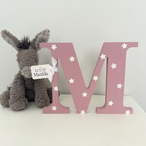Blush pink wooden letter M with white star pattern. The letter is decorated with a white tag that says 'is for Matilda'.
