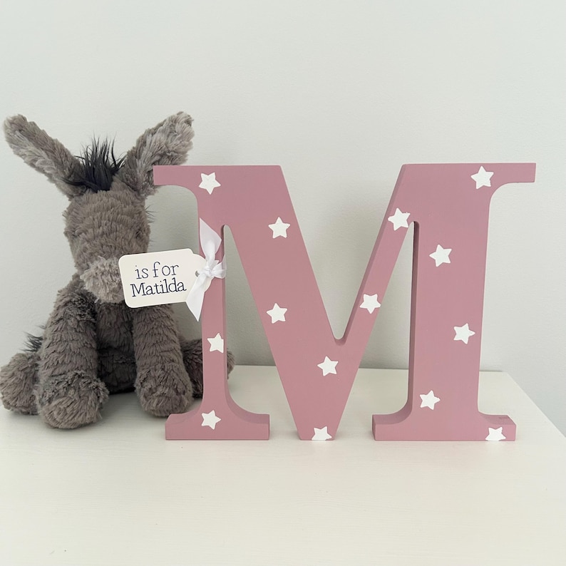 Blush pink wooden letter M with white star pattern. The letter is decorated with a white tag that says 'is for Matilda'.