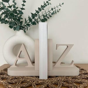 A pair of painted wooden bookends in the colour Oat, shaped as the letters 'A' and 'Z', supporting a book, with greenery visible in the background, all on a woven surface