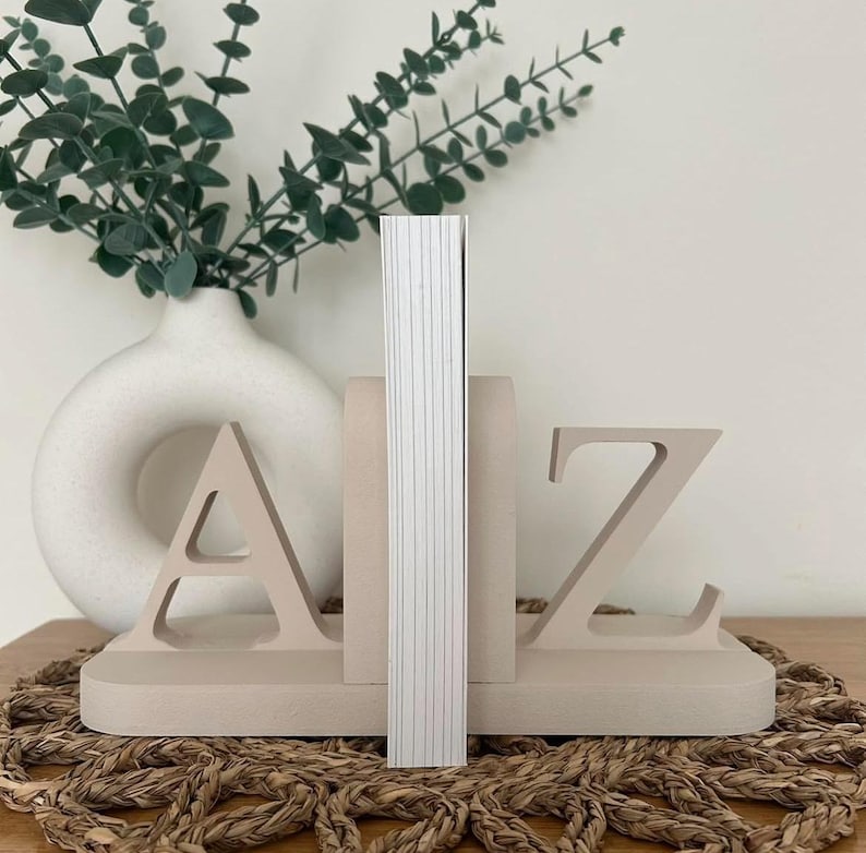 A pair of painted wooden bookends in the colour Oat, shaped as the letters 'A' and 'Z', supporting a book, with greenery visible in the background, all on a woven surface