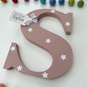 Pode incluir: Uma grande letra "S" de madeira pintada em rosa suave, adornada com estrelas brancas. Uma etiqueta amarrada com uma fita branca diz "is for Sophia". A letra faz parte de um conjunto decorativo, ideal para um quarto infantil.