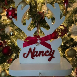 May include: A white wooden Christmas stocking holder shaped like reindeer antlers, with a red ribbon and the name "Nancy" in red glitter. A small bell and ornament hang from the antlers. The holder is displayed in front of a decorated Christmas tree.