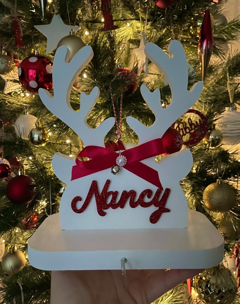 May include: A white wooden Christmas stocking holder shaped like reindeer antlers, with a red ribbon and the name "Nancy" in red glitter. A small bell and ornament hang from the antlers. The holder is displayed in front of a decorated Christmas tree.