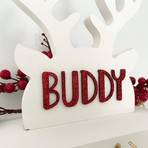 May include: White wooden reindeer decoration with red glitter text that reads "BUDDY". The reindeer has antlers and is decorated with red berries. A festive holiday decoration.