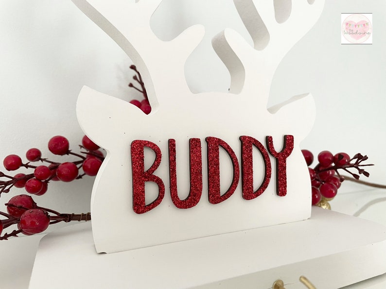 May include: White wooden reindeer decoration with red glitter text that reads "BUDDY". The reindeer has antlers and is decorated with red berries. A festive holiday decoration.