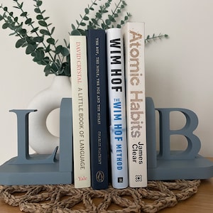 A pair of denim blue painted wooden bookends, shaped as the letters 'L' and 'B', supporting a row of books, with a white vase containing artificial greenery visible in the background, all on a woven surface