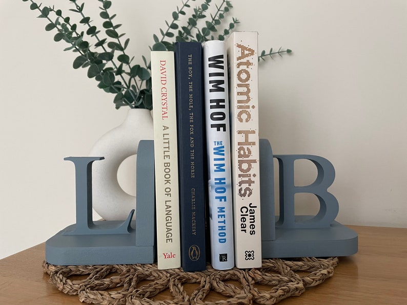 A pair of denim blue painted wooden bookends, shaped as the letters 'L' and 'B', supporting a row of books, with a white vase containing artificial greenery visible in the background, all on a woven surface