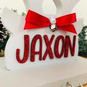 May include: A white wooden reindeer head with the name "JAXON" in red glitter letters. A red satin bow with a small gold bell and pearl accents adorns the top. Festive holiday decor.