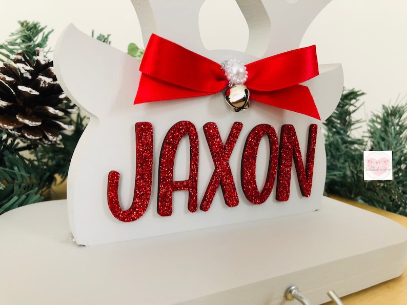 May include: A white wooden reindeer head with the name "JAXON" in red glitter letters. A red satin bow with a small gold bell and pearl accents adorns the top. Festive holiday decor.