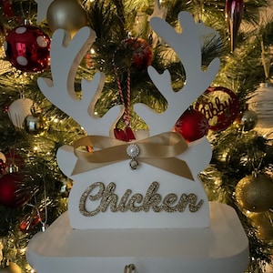 May include: White wooden reindeer Christmas ornament with gold glitter text that reads "Chicken". The ornament has a gold ribbon and pearl embellishment. The background is a Christmas tree with red and gold ornaments.