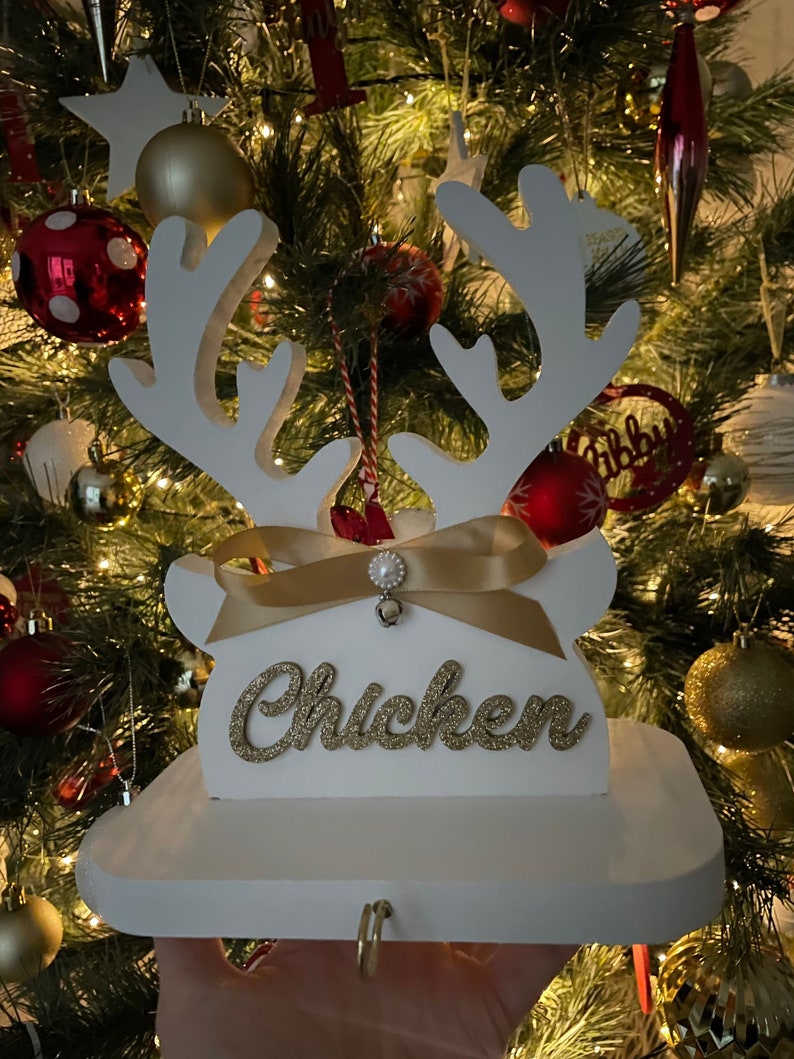 May include: White wooden reindeer Christmas ornament with gold glitter text that reads "Chicken". The ornament has a gold ribbon and pearl embellishment. The background is a Christmas tree with red and gold ornaments.
