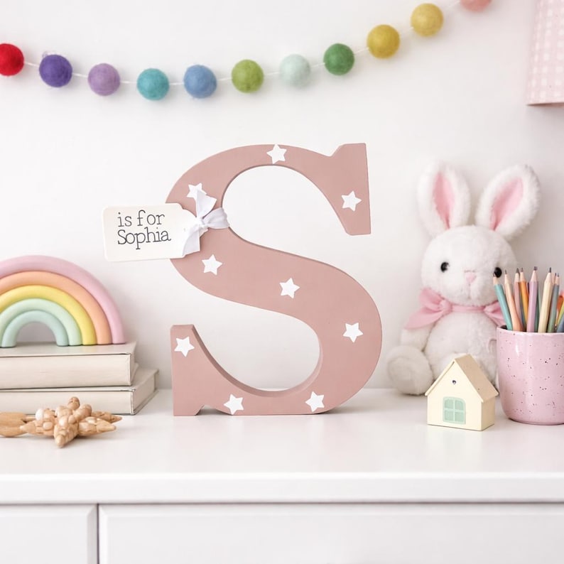 a painted wooden letter S, in dusky pink white white stars; with a white tag on the top left hand side of letter, attached with white ribbon and the name Sophia on. the freestanding letter is styled on a dresser in a girls bedroom