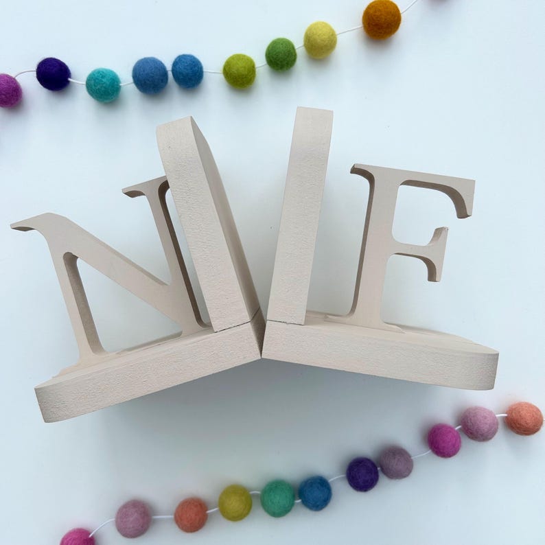 a pair of letter bookends, N & F, painted in a natural oat colour, on a white background with two strings of coloured felt balls used as a photo prop