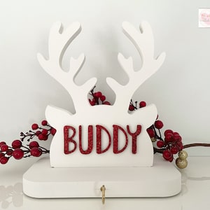 May include: A white wooden reindeer head with antlers and the word "BUDDY" in red glitter. The reindeer is decorated with red berries and a small gold-colored keyhole. A festive holiday decoration.