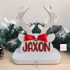 May include: A white wooden reindeer head Christmas stocking holder with the name "JAXON" in red glitter letters. It features a red bow, a small silver bell, and a hook for hanging stockings. Festive holiday decor.