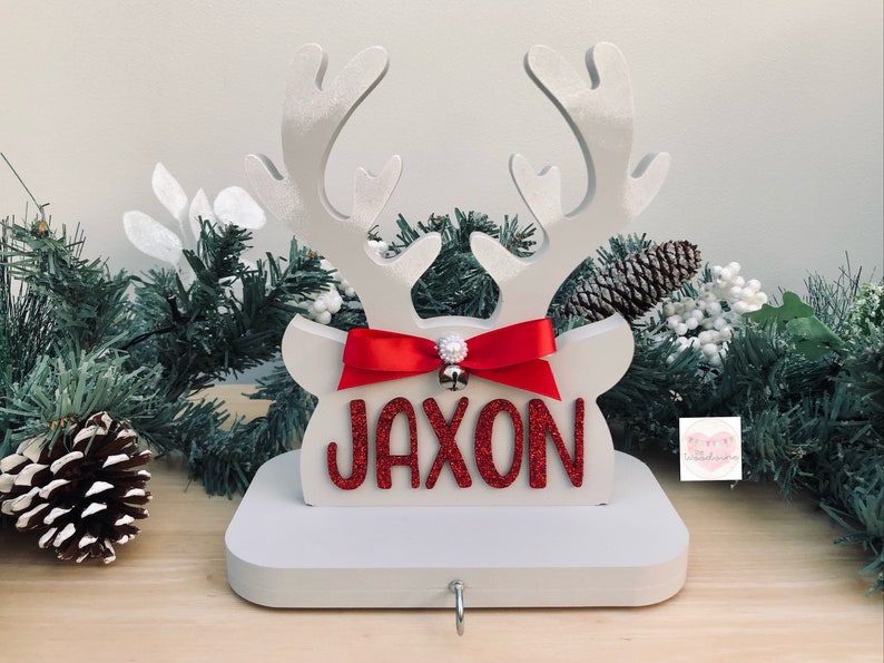 May include: A white wooden reindeer head Christmas stocking holder with the name "JAXON" in red glitter letters. It features a red bow, a small silver bell, and a hook for hanging stockings. Festive holiday decor.