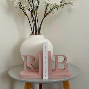 A pair of dusky pink painted wooden bookends, shaped as the letters 'R' and 'B', supporting a book, with a white vase containing artificial greenery visible in the background, all on a small side table.
