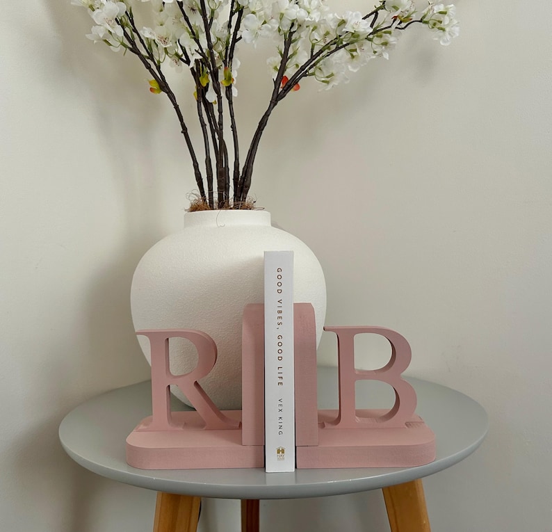 A pair of dusky pink painted wooden bookends, shaped as the letters 'R' and 'B', supporting a book, with a white vase containing artificial greenery visible in the background, all on a small side table.