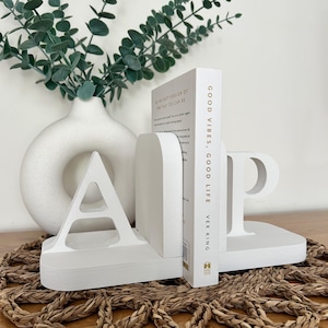 A pair of white painted wooden bookends, shaped as the letters 'A' and 'P', supporting a book, with a white vase containing artificial greenery visible in the background, all on a woven surface