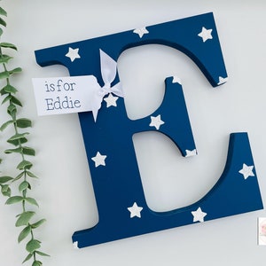 Pode incluir: Uma letra "E" de madeira azul marinho decorada com estrelas brancas. Uma pequena etiqueta amarrada com uma fita branca diz "is for Eddie". A letra &eacute; um item decorativo, possivelmente para um quarto infantil.