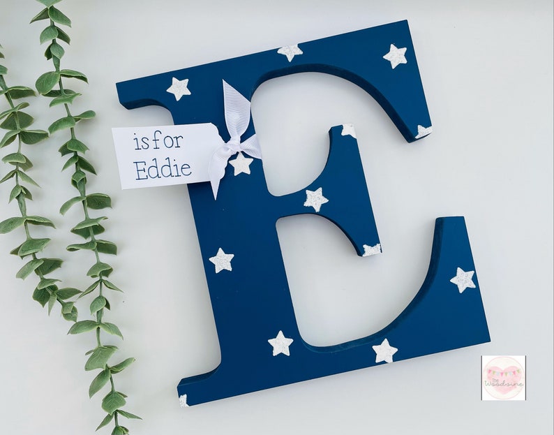 Pode incluir: Uma letra "E" de madeira azul marinho decorada com estrelas brancas. Uma pequena etiqueta amarrada com uma fita branca diz "is for Eddie". A letra &eacute; um item decorativo, possivelmente para um quarto infantil.