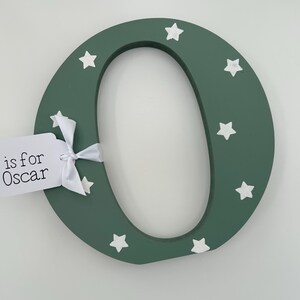 Pode incluir: Uma letra "O" de madeira verde escuro decorada com estrelas brancas. Uma etiqueta branca amarrada com uma fita branca diz "is for Oscar". A letra &eacute; um item decorativo, possivelmente para um quarto infantil.