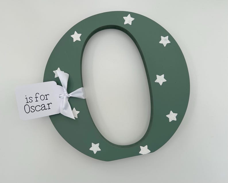 Pode incluir: Uma letra "O" de madeira verde escuro decorada com estrelas brancas. Uma etiqueta branca amarrada com uma fita branca diz "is for Oscar". A letra &eacute; um item decorativo, possivelmente para um quarto infantil.