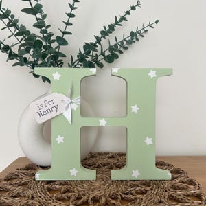 freestanding, bright green initial H with white stars on and a white tag attached to the left with a white ribbon with the words “is for Henry” on.